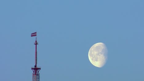 Latvian-flag-flies-in-the-TV-tower,-the-moon-flies-in-the-sky-at-sunset