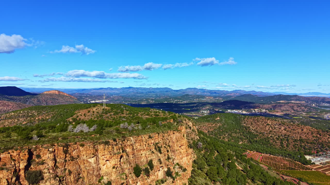 Vista desde una montaña con vistas a valles y campos, con cielos azules y nubes dispersas durante el día en una zona rural.