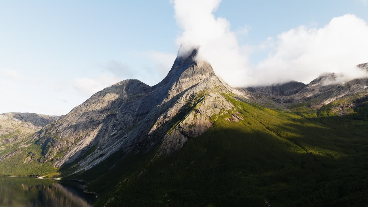 el pico de la montaña estéril con pendiente de hierba domina el lago mientras las nubes se reúnen en la parte superior, stetind noruega