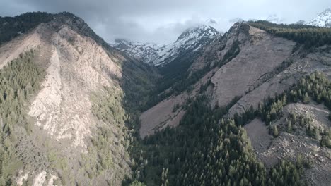 Panning-up-shot-of-summer-mountains-at-low-altitude-and-snowy-cloudy-peaks-near-the-top,-shot-in-Big-Cottonwood-Canyon,-Utah-with-sunset-lighting-coming-in-from-the-right-side