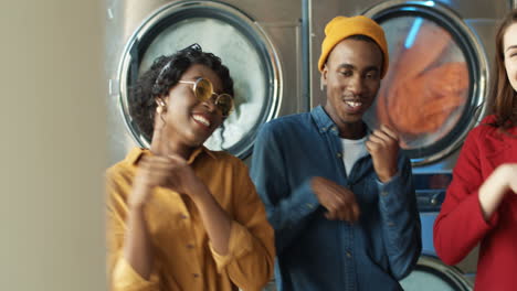 young mixed races cheerful stylish girls and guy having fun and dancing in laundry service room while machines washing on background