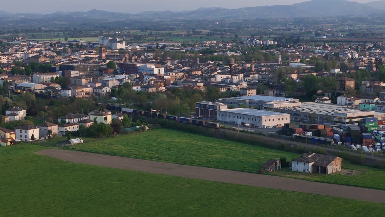 Treno merci che passa per Fiorenzuola d’Arda, Piacenza, Emilia Romagna, Italia, sulla linea ferroviaria Milano-Bologna con terreni agricoli della Pianura Padana e la lontana cresta degli Appennini settentrionali. Ripresa in movimento con drone