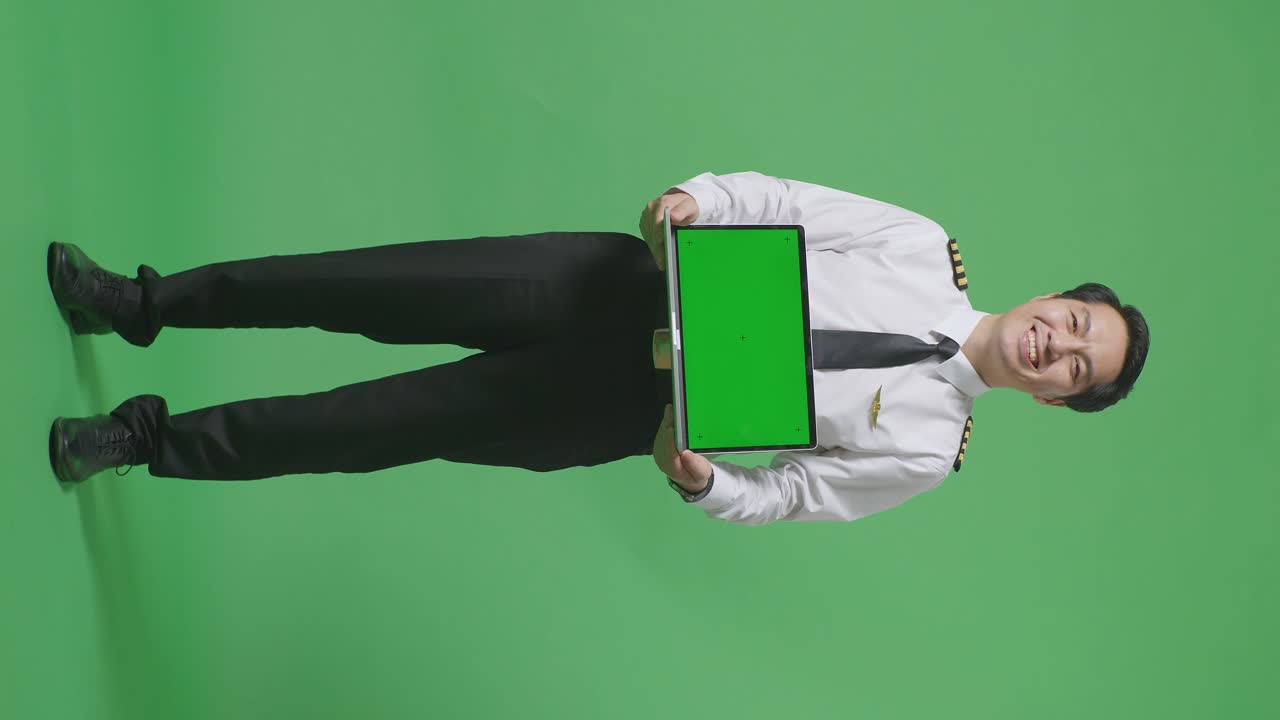 cuerpo lleno de piloto hombre asiático sonriendo y mostrando pantalla verde portátil a la cámara mientras estaba de pie en el estudio de fondo pantalla verde