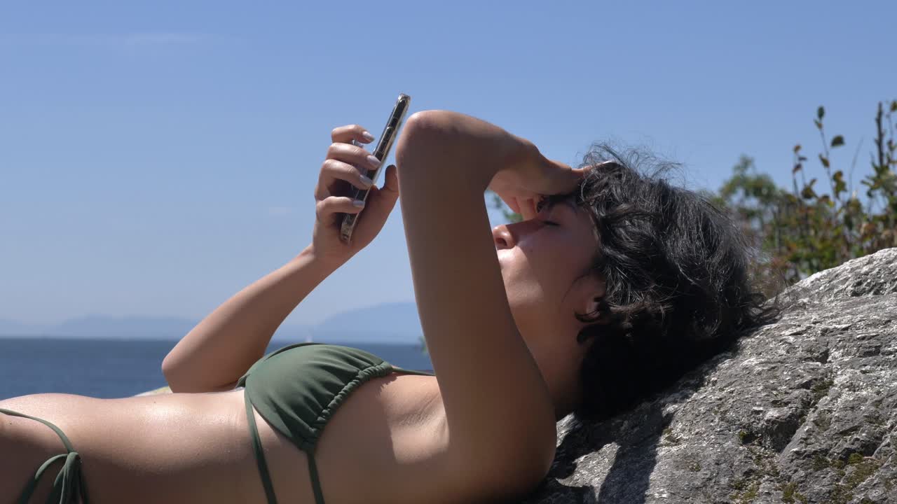 chica morena en bikini tomando el sol y usando un teléfono inteligente mirando a la cámara