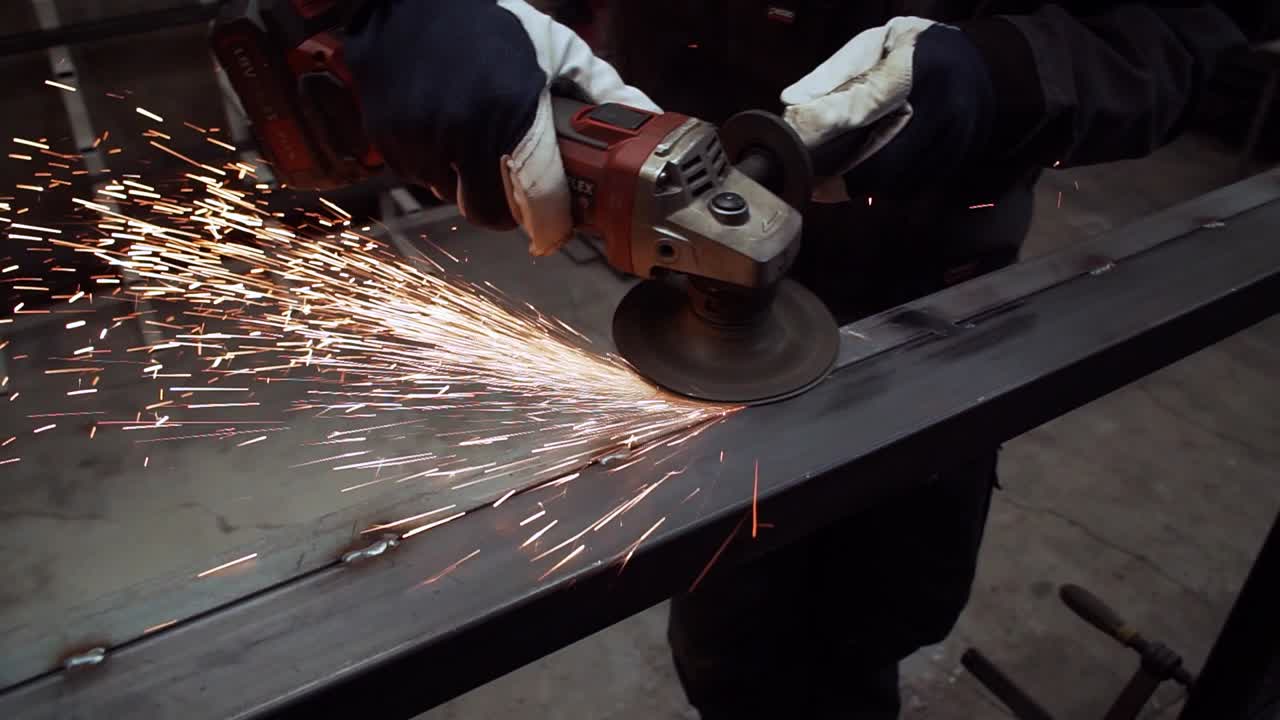 The worker is grinding metal beam with the angle grinder, there are a lot of sparks. Camera rotates clockwise