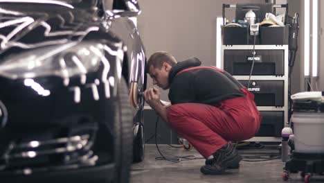 Polishing-car-with-polish-mashine.-Worker-in-red-suit-cleaning-a-black-expensive-car.