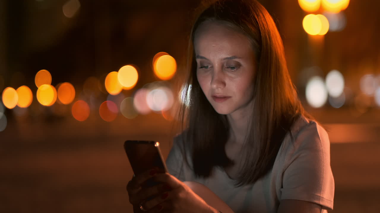 A young woman looks into the smartphone and writes text messages on the Internet against the backdrop of the night city. Girl businessman working on vacation remote work via mobile phone. Gadget addiction