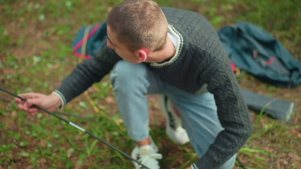 Closeup partial view of man squatting while arranging tent poles on forest floor during camping setup, surrounded by dry leaves, pine needles, and scattered gear, with blurred companion nearby