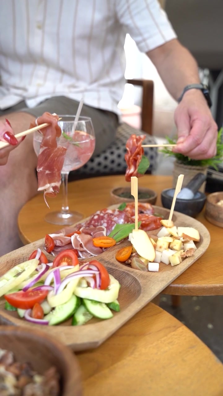 Un tagliere di salumi e formaggi con una varietà di affettati, formaggi a fette e insalata fresca con cetrioli, pomodori e cipolle rosse. Si vedono mani che prendono gli alimenti.
