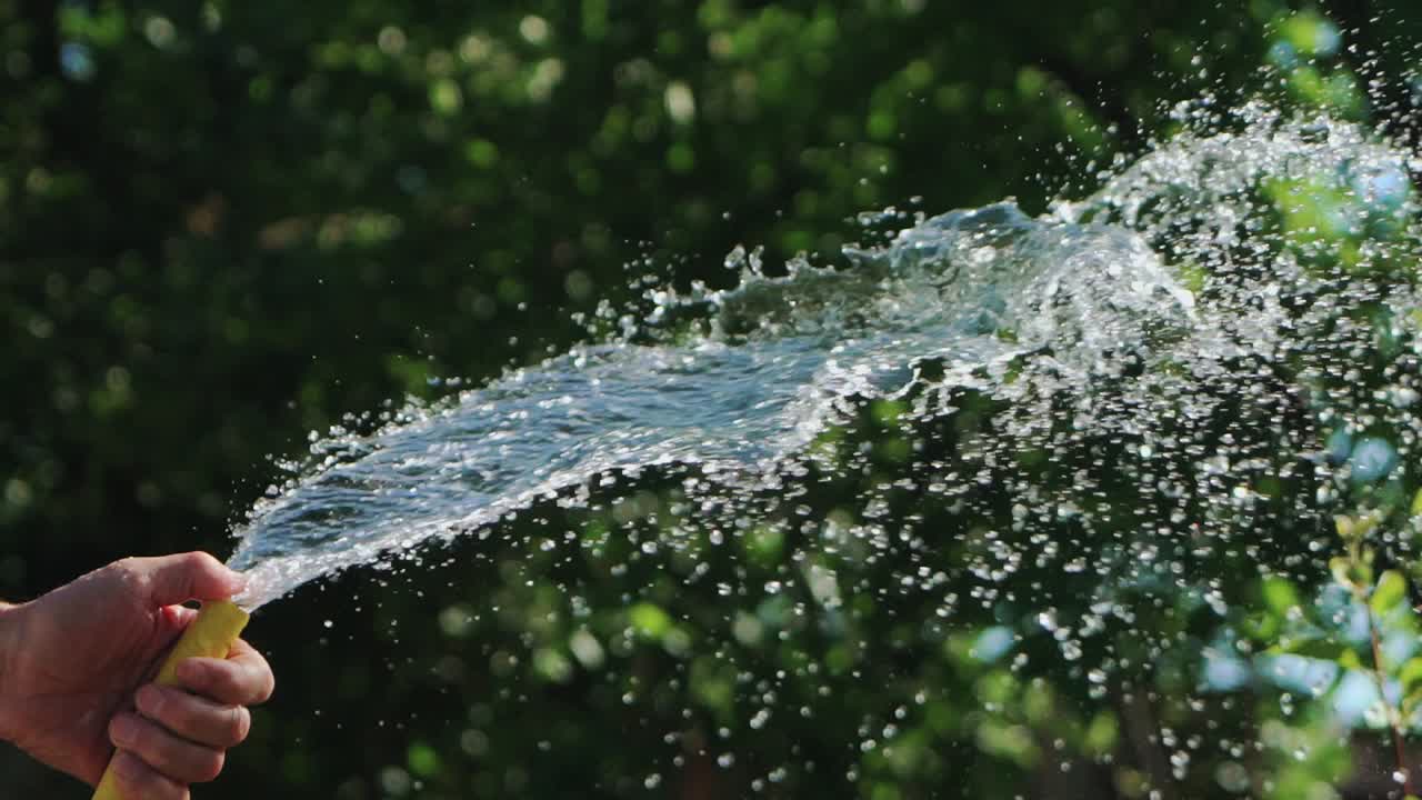 A man with a yellow hose in his hands directs a stream of water directly and higher regulating the process of watering plants in the yard. Close-up.