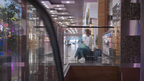 wide view of a young woman seated in a modern shopping mall, sliding through her phone with decorative lighting and a festive atmosphere in the background