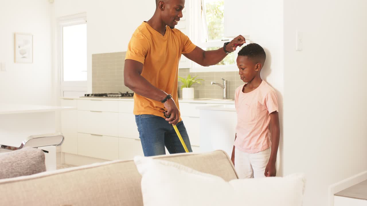 African American father measuring son's height at wall at home holding tape measure tracking growth