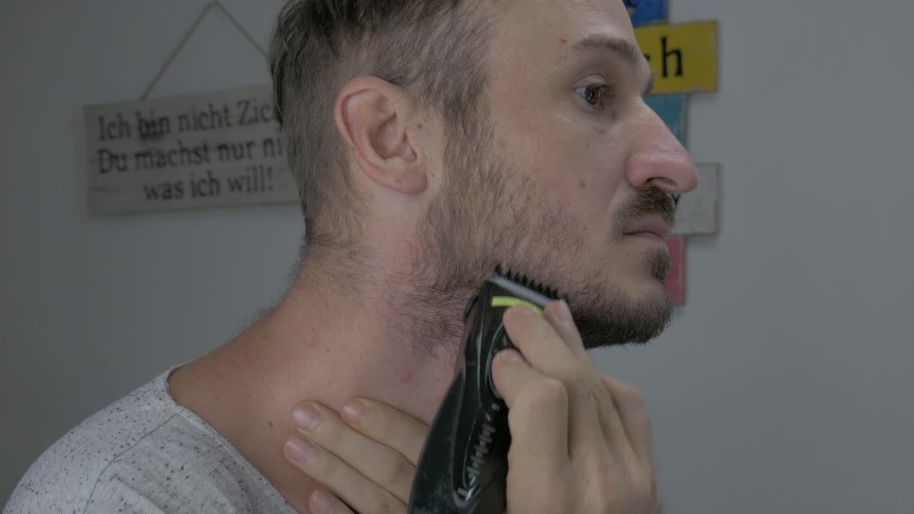 A Caucasian male is trimming his facial hair with an electric shaver to look gentle, The close up footage shows a man's face while he is trimming and shaving his beard