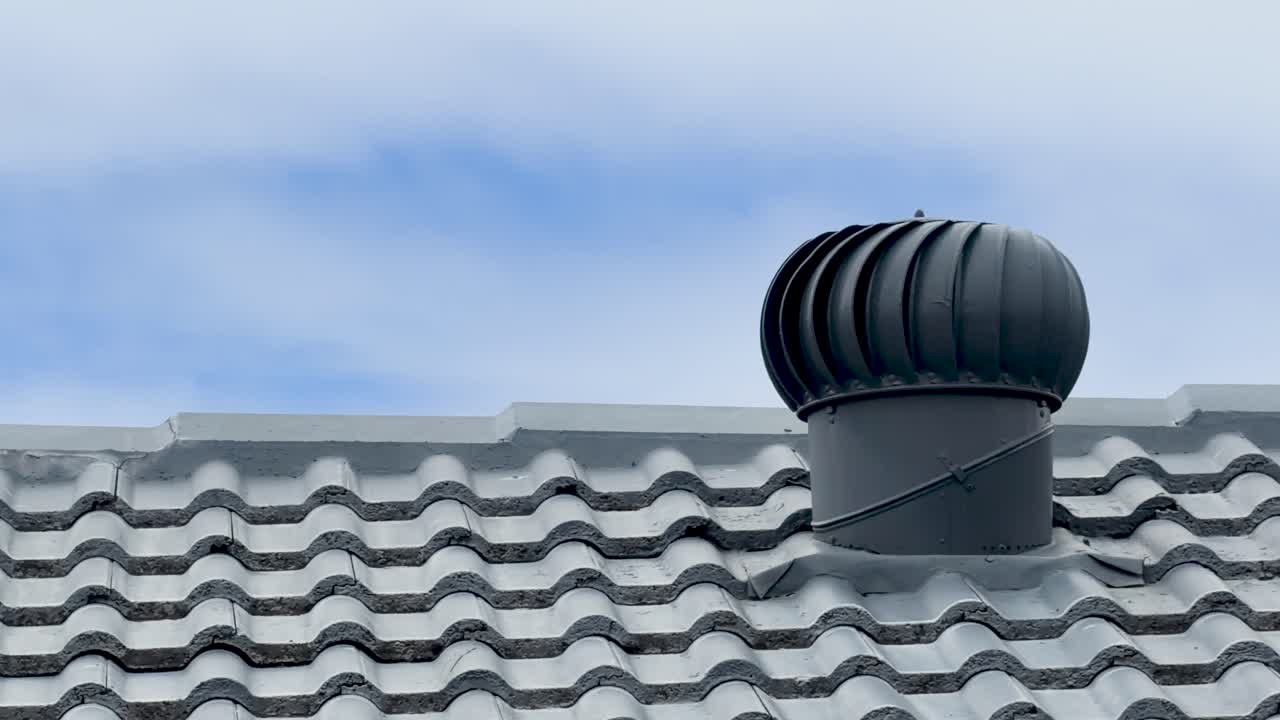 A dark metal turbine vent rotates on a gray tiled roof under blue skies