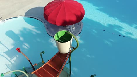 antena ascendente revela parque acuático en la piscina, sombrillas rojas salpican un día soleado, sin gente