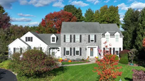 Suburban-home-in-America-with-grey-stone,-white-siding,-colorful-autumn-trees,-American-flag,-and-Halloween-decor