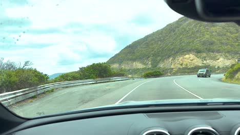 a car navigates a winding coastal road with ocean views and lush hills under bright daylight