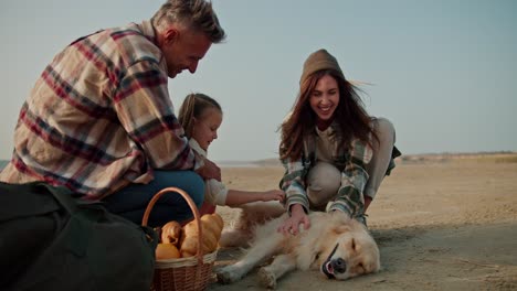 A-happy-brunette-girl-in-a-hat-in-a-green-checkered-shirt,-together-with-her-husband,-strokes-a-large-cream-colored-dog-and-near-them-their-little-daughter-sits-and-relaxes-during-their-picnic-on-the-deserted-seashore-in-the-summer