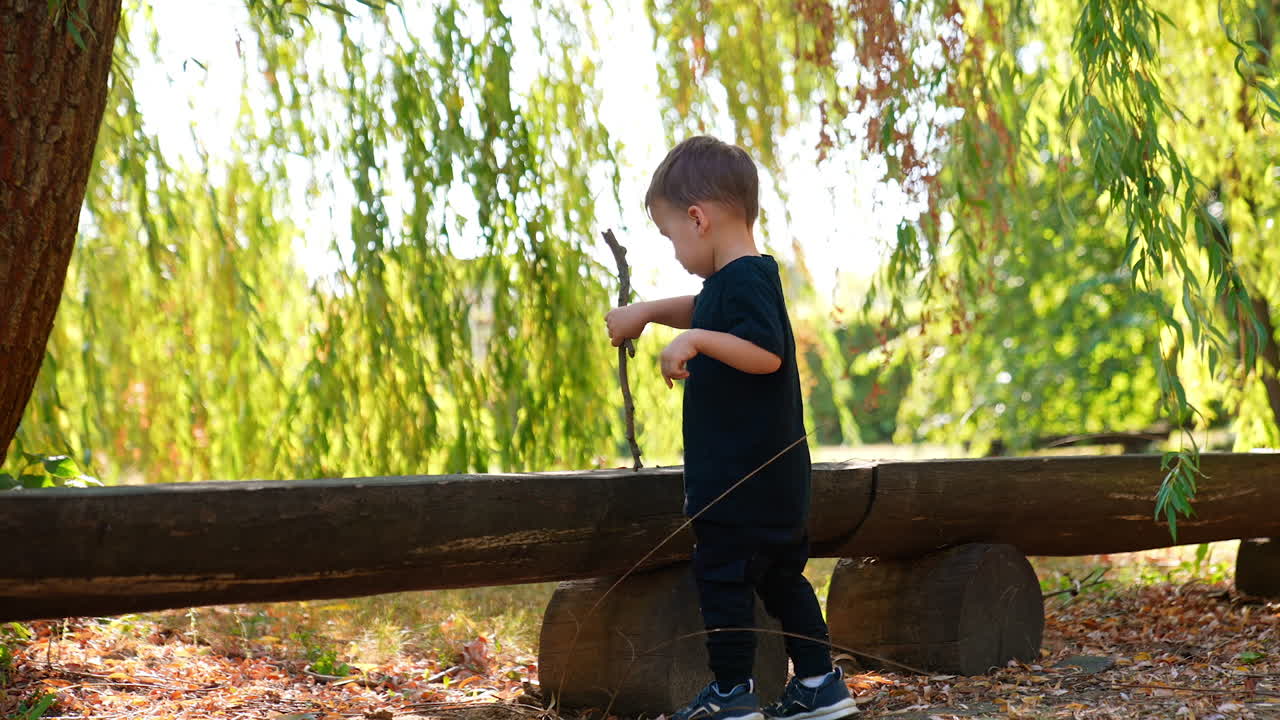 Cute Caucasian baby boy wearing dark clothes stands at the long wooden bench outdoors. Active toddler taps the bench with a stick.
