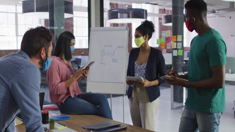 Mixed-race-business-colleagues-wearing-face-masks-having-a-discussion-in-meeting-room-making-notes