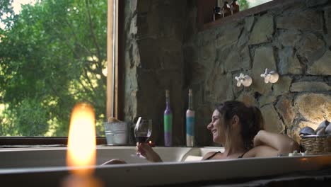 young attractive female having a fancy bath in a jacuzzi drinking wine looking by big green nature window able to slow motion 60fps