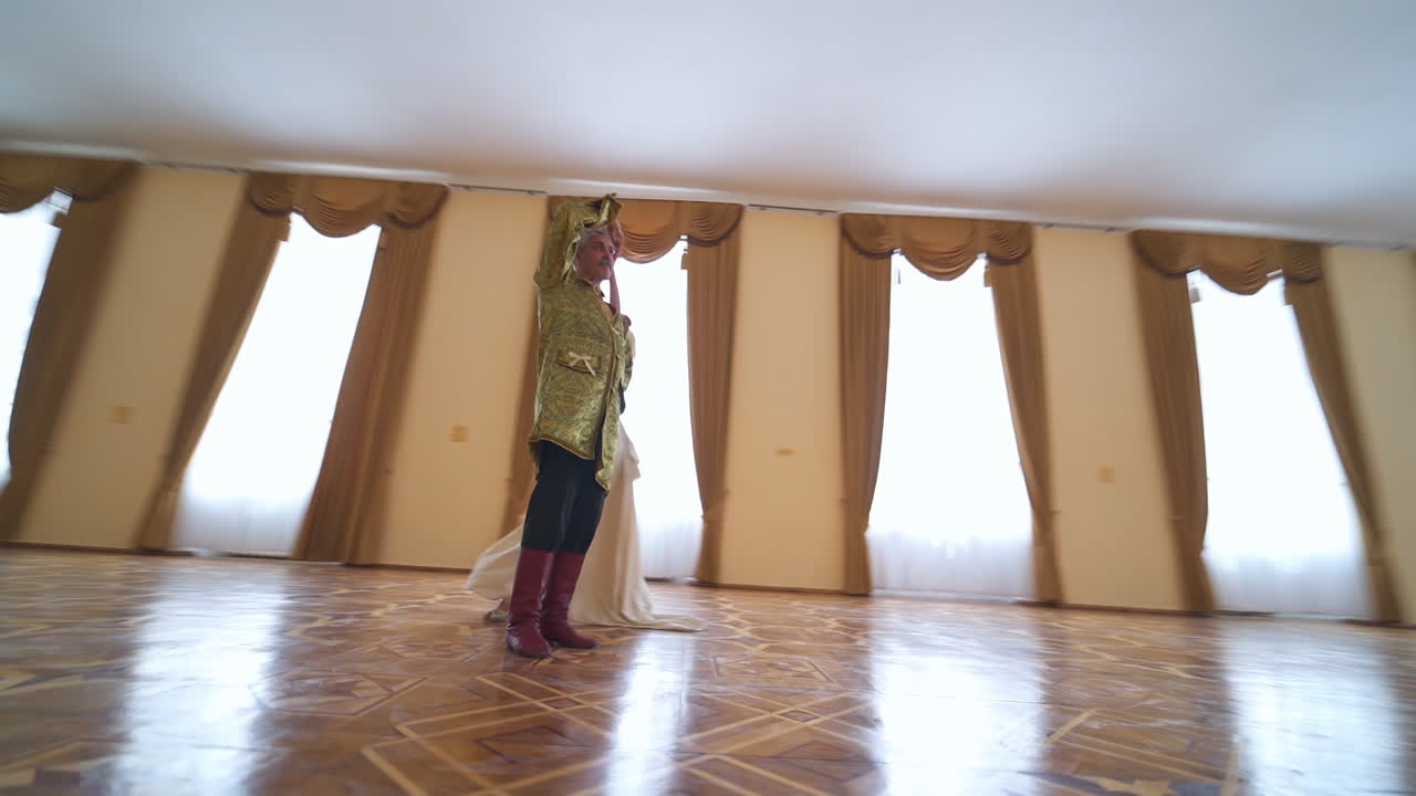 Adult lady in long white historic dress circling around the man in golden jacket. Couple performing old kind of dance in the spacious hall with parquet floor. Low angle view.