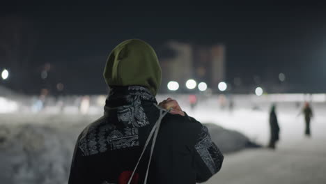back view of skater wearing hooded jacket walking away with skate boot over shoulder as visible cold breath escapes into night air under distant lights on snowy path with people in background