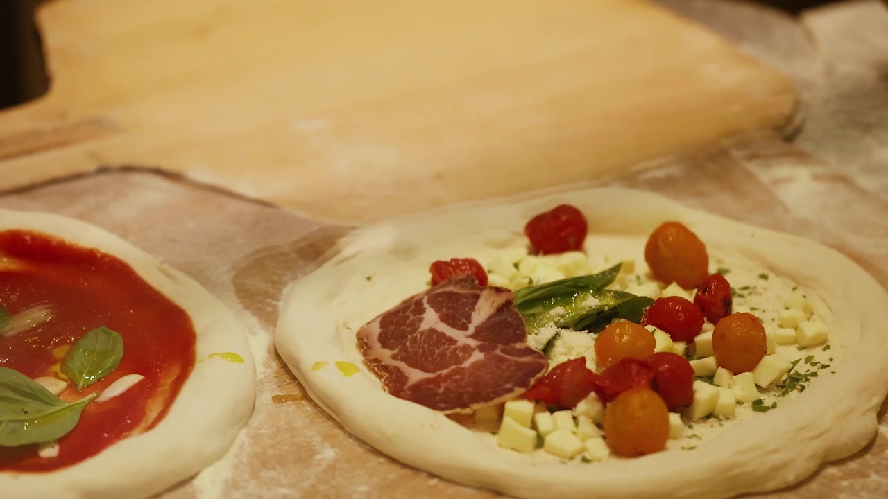 Primo piano di mani che guarniscono l'impasto crudo della pizza con prosciutto, basilico e pomodorini.