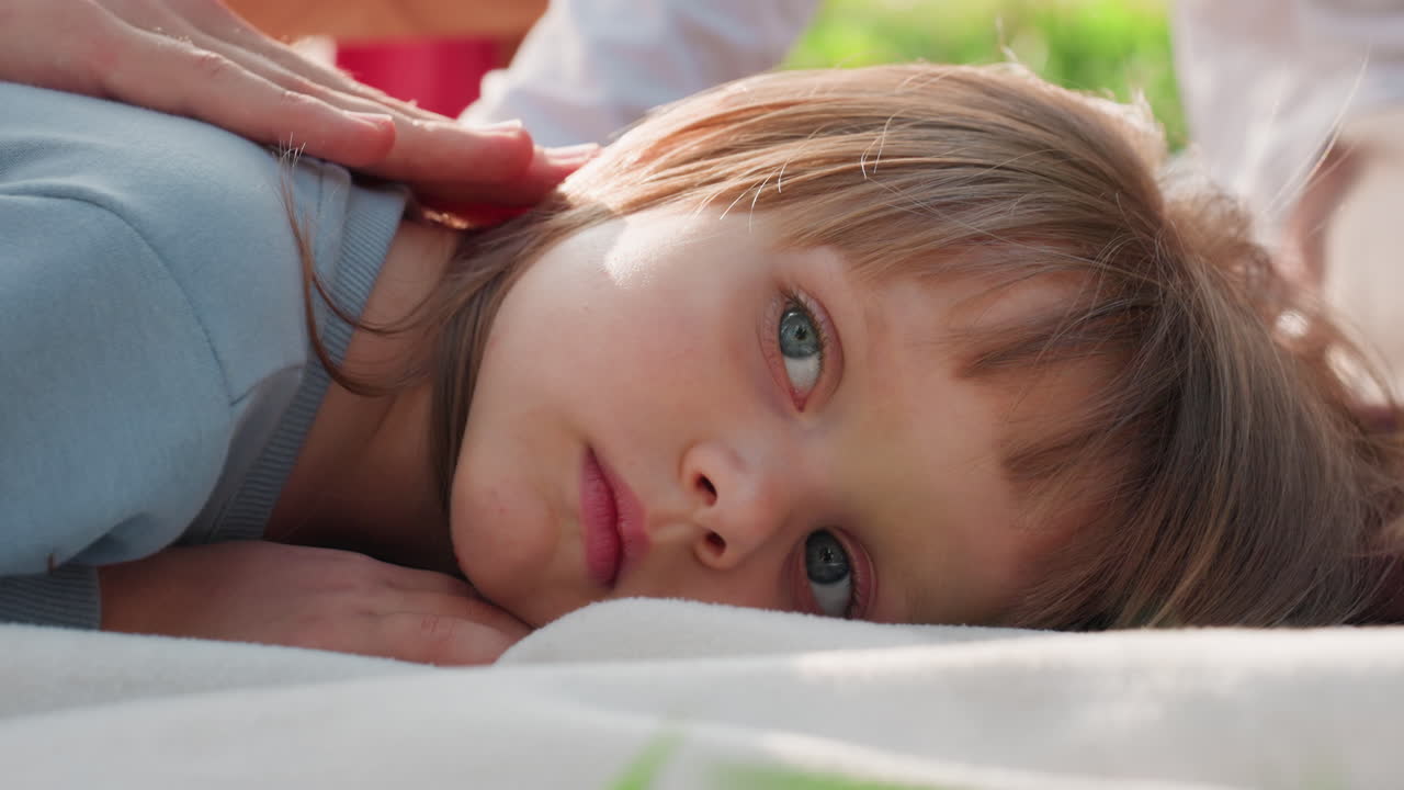 Close up of exhausted young girl lying quietly on blanket while sibling gently rubs her head to comfort her, sunlight softly highlighting her peaceful face, showing emotion,and family warmth