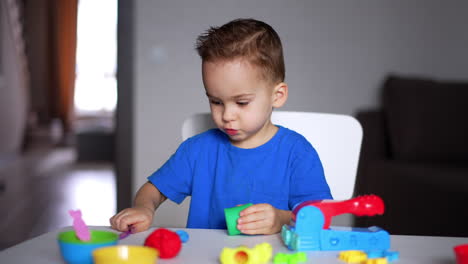 beautiful caucasian toddler wearing blue t-shirt sits at desk. kid tries to cut the piece of plasticine with a stick.