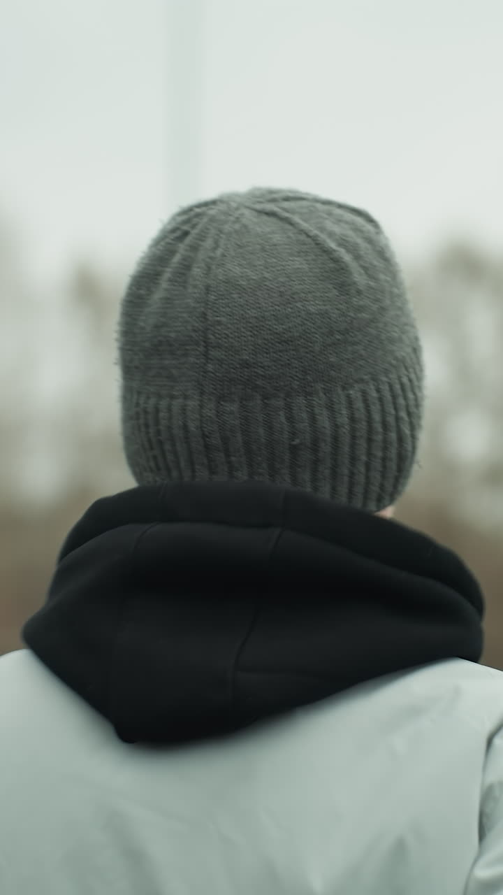 Back view of a person jogging on a track, wearing a gray jacket and a black beanie, with a blurred view of a woman in a brown jacket and another person jogging opposite each other
