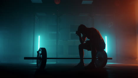 exhausted and depressed sportsman in dark gym sitting near barbell and resting dramatic silhouette