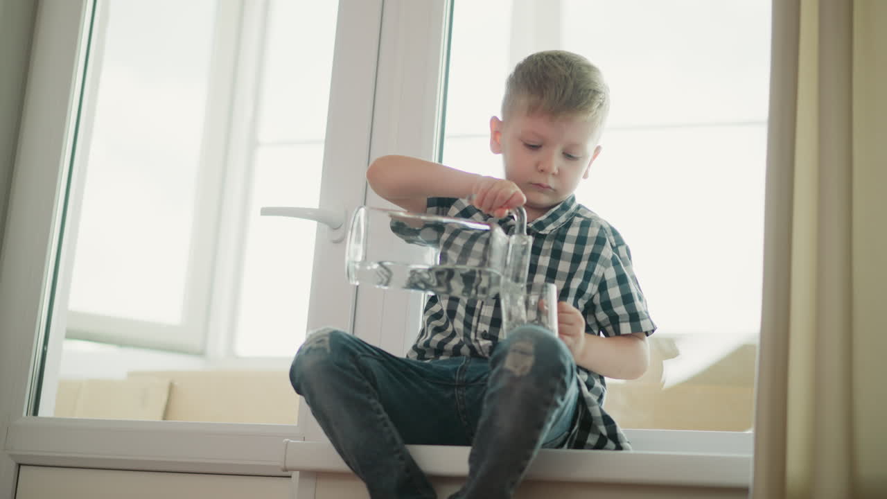Young boy in plaid shirt sitting near window carefully turning water from glass jar in right hand into cup in left hand while focusing on pouring with natural daylight filling bright room interior