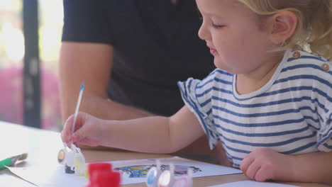 Close-up-of-father-and-young-daughter-having-fun-at-home-sitting-at-table-and-painting-decoration-together---shot-in-slow-motion