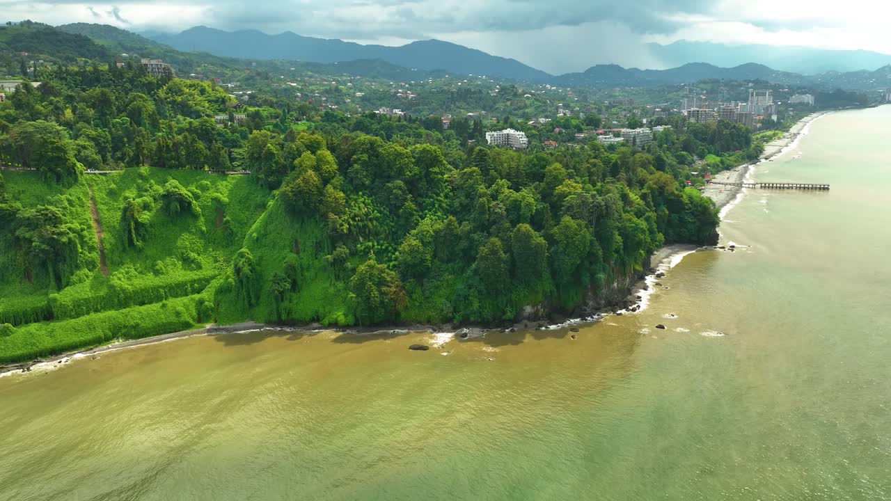 Aerial view of a lush green coastal area where rolling hills meet the brownish waves of the sea, with distant mountains and soft sunlight highlighting the natural landscape