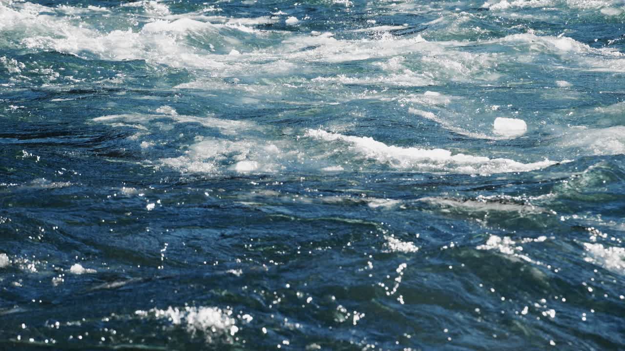 Rushing river water sparkles in the sunlight, creating mesmerizing patterns and waves. The vibrant blue water energizes the surrounding environment, showcasing nature's beauty at noon