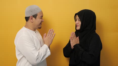 happy asian muslim couple doing greeting gesture, apologize and forgiving each other on eid mubarak tradition, against yellow