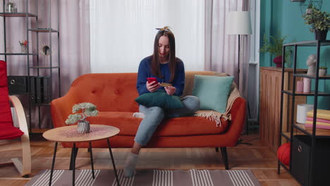 Woman-sitting-at-home-using-credit-bank-card-and-smartphone-while-transferring-money-online-shopping