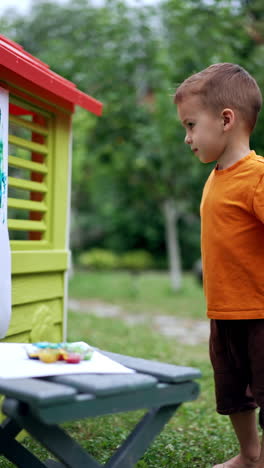 beautiful baby boy using bright paints to create a picture. cute kid painting in the garden and looks at his work with satisfaction. vertical video.