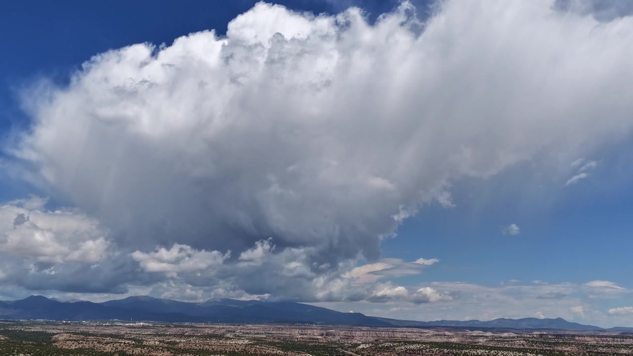 4K UHD drone timelapse capturing a massive, billowing cumulus cloud morphing and expanding over rugged desert mesa formations. The towering white cloud dominates the blue sky above the arid landscape.