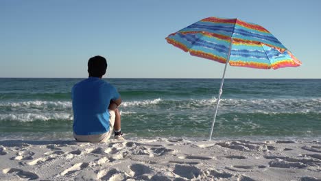 medium steady shot of a man wearing a blue t-shirt sitting under a colorful sunshade umbrella on the beach, gazing at the sea and horizon from behind with a clear blue sky in the background