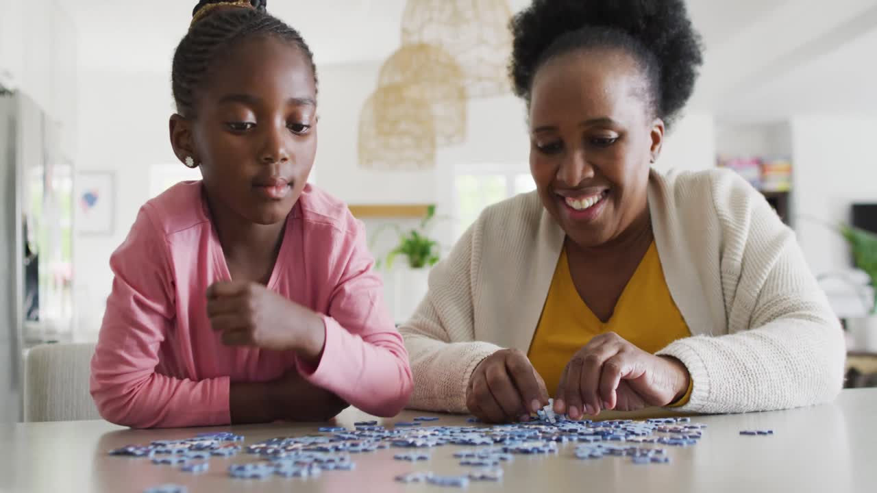 Happy african american granddaughter and grandmother doing jigsaw puzzle at home, copy space