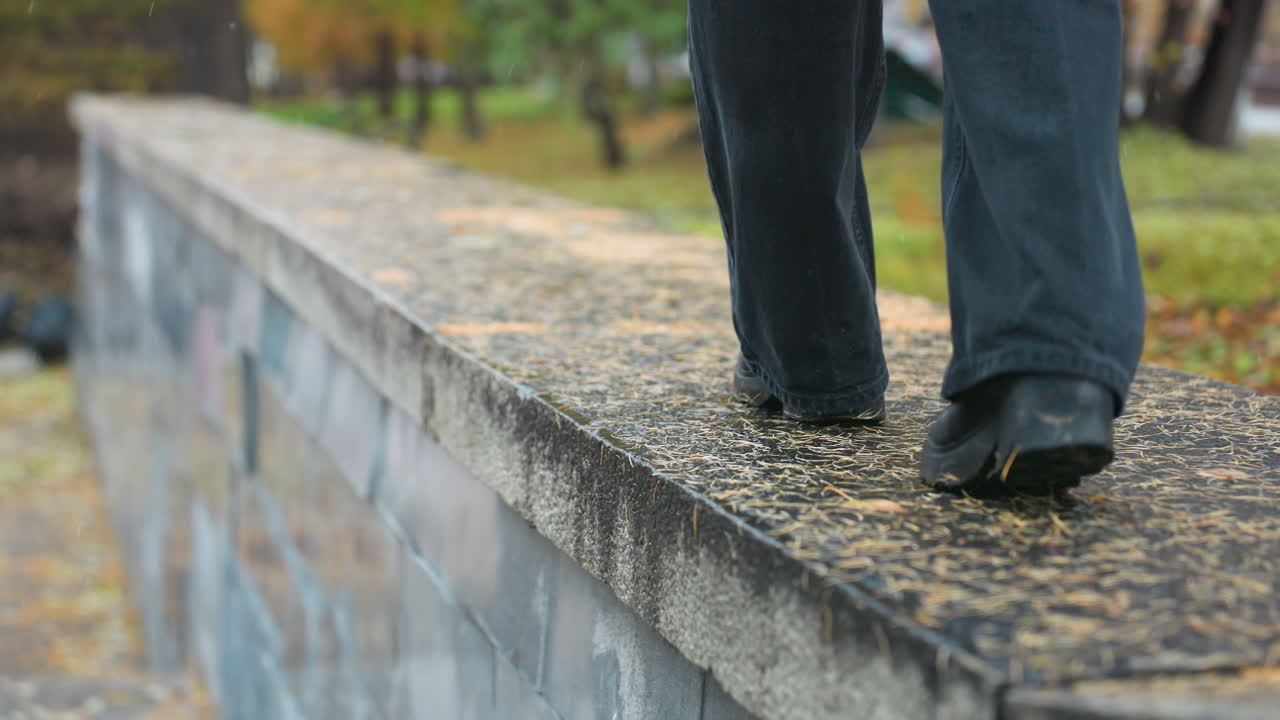 Back view of person wearing black trousers and boots carefully walking along wide wet stone path during rainy autumn day, with fallen pine needles scattered on ground and blurred park backdrop