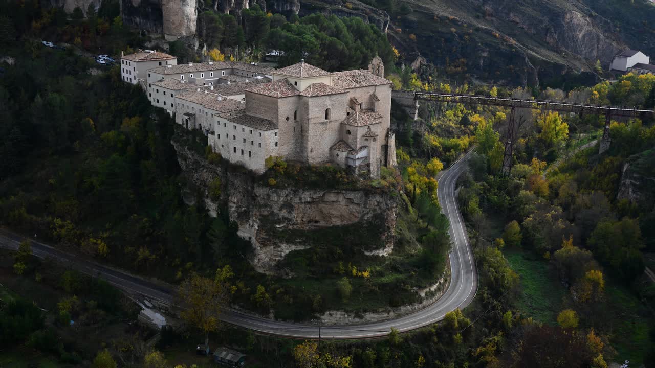 A road winds through the deep Huecar gorge in Cuenca, Spain, passing below the historic Parador de Cuenca, a former convent perched on the cliffs above.