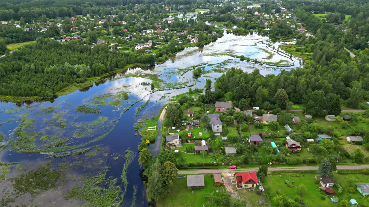 Drone panoramic dolly above flooded river that rose to edge of homes, cloudy blue sky reflects in water, neighborhoods between forest tree stands