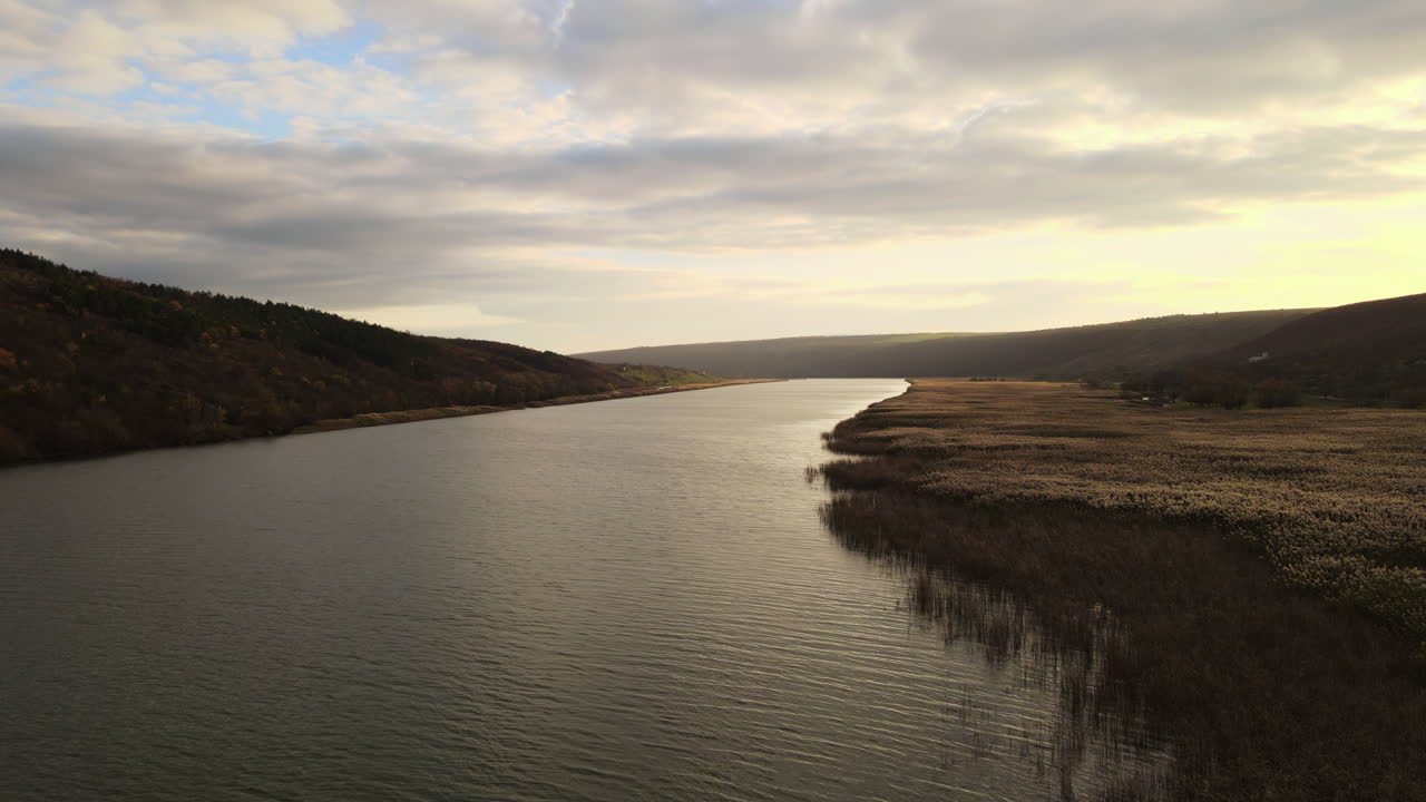 A stunning sunset casts warm hues on the calm river, reflecting the sky. Hills and lush grass provide a scenic setting in the peaceful countryside