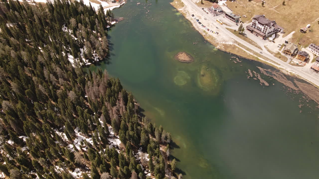 Camera tilts upward from an emerald lake and mountain resort, revealing a vast green valley surrounded by dense forest and dramatic snow‑capped Dolomite mountains