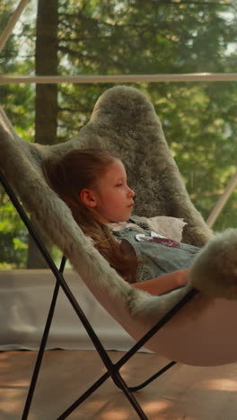 calm child sitting in fur armchair relaxes during hot day while watching landscapes of mountain forest. preschooler girl in glamping hides from heat outdoors