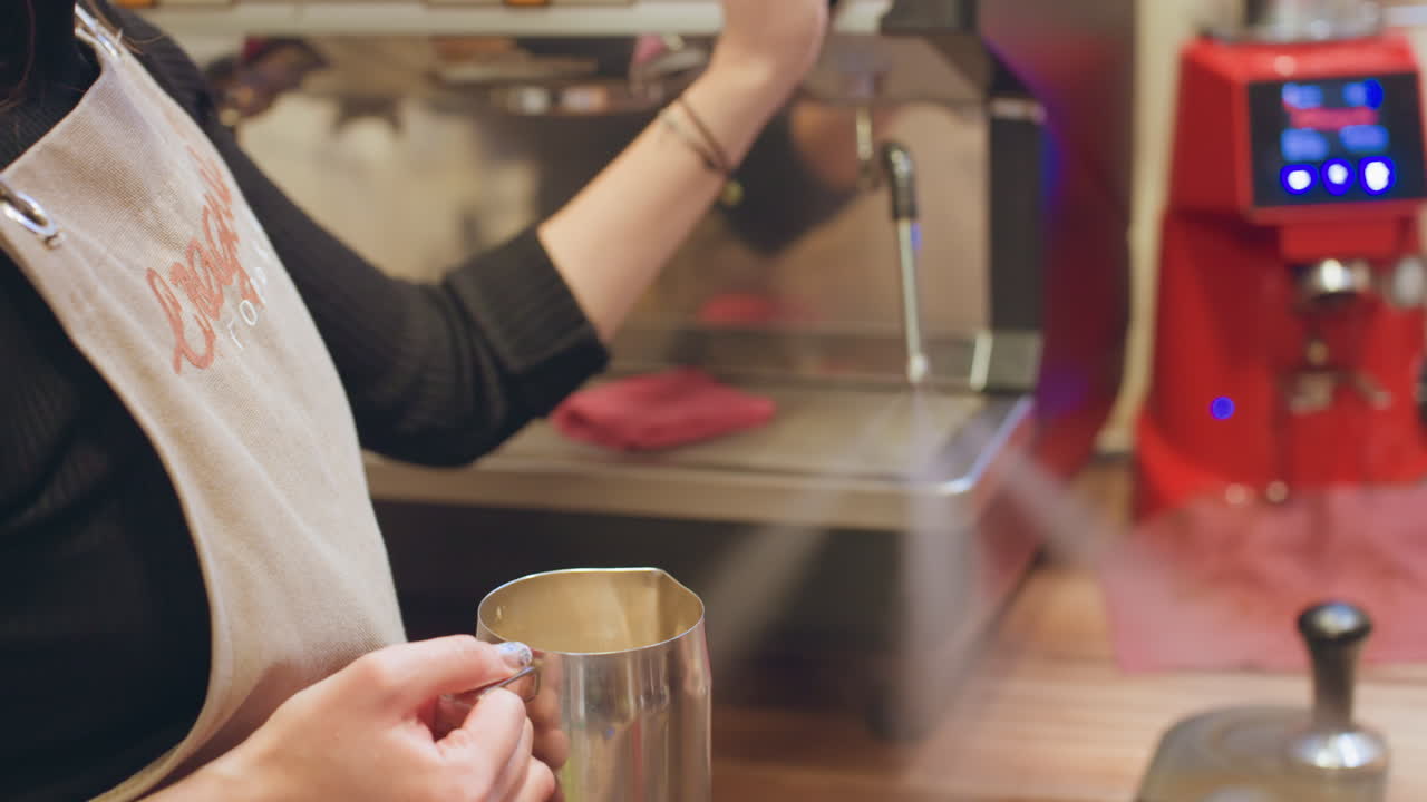 Barista donna che monta il latte con la macchina da caffè espresso, mani che tengono il bricco in acciaio inossidabile e regolano la lancia vapore, bancone caldo, dettagli del grembiule, primo piano sulla texture della schiuma e sulla crema, intimo caffè mattutino.