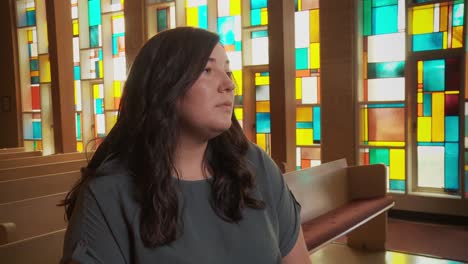 Close-up-of-a-pretty-young-ethnic-female-staring-sadly-at-the-altar-inside-a-church-sanctuary-while-sitting-in-a-pew